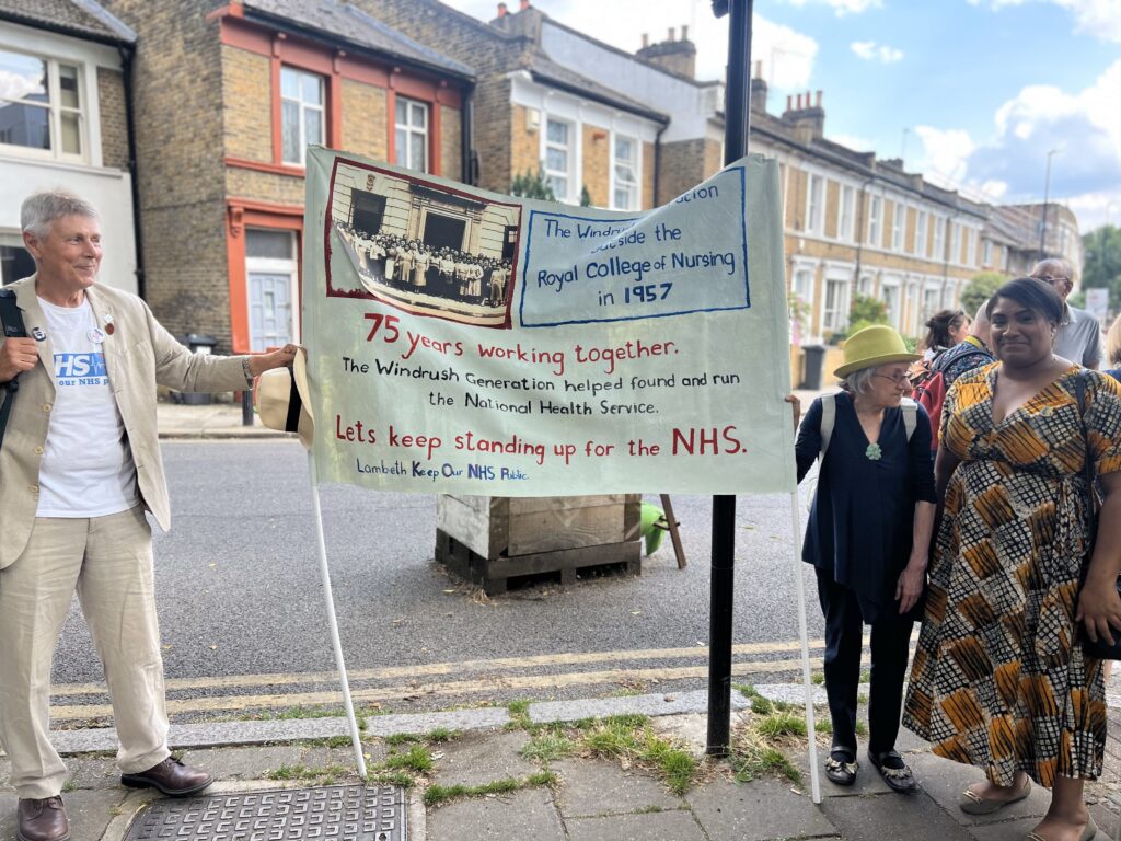 Bell stands by a banner with local Keep our NHS Public activists acknowledging the Windrush Generation's contribution to building our NHS.