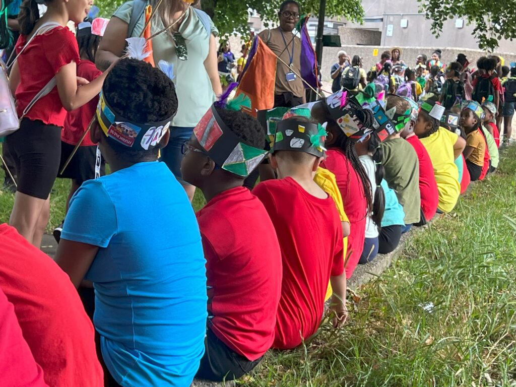 Local school pupils sit wearing headdresses with Caribbean themes and colours.
