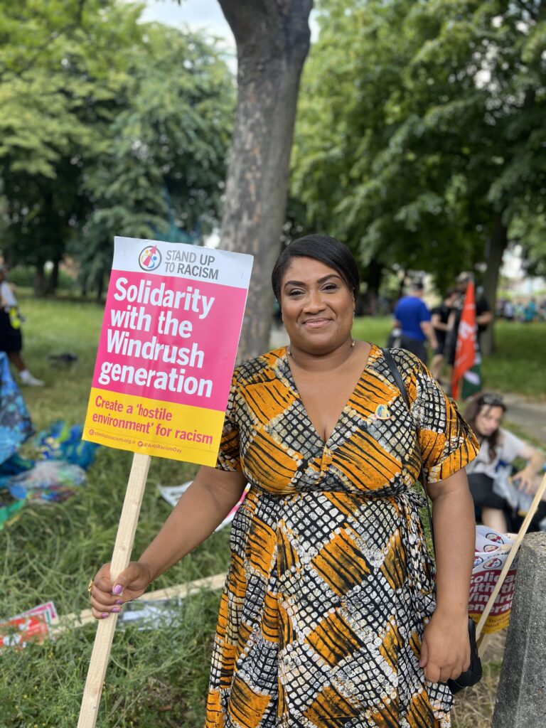 Bell stands holding a sign that says 'Solidarity with the Windrush Generation: create a "hostile environment" for racism.'