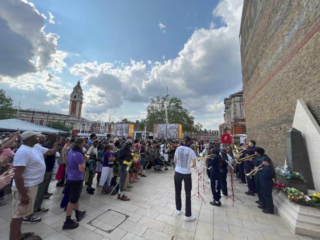A large crowd gathers around trumpet players in Windrush Square in front of the African and Caribbean War Memorial, overlooked by the clocktower of Lambeth Town Hall.