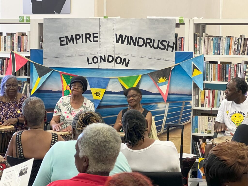 Windrush elders drumming in front of a mocked up Empire Windrush frontage with Caribbean flags attached.