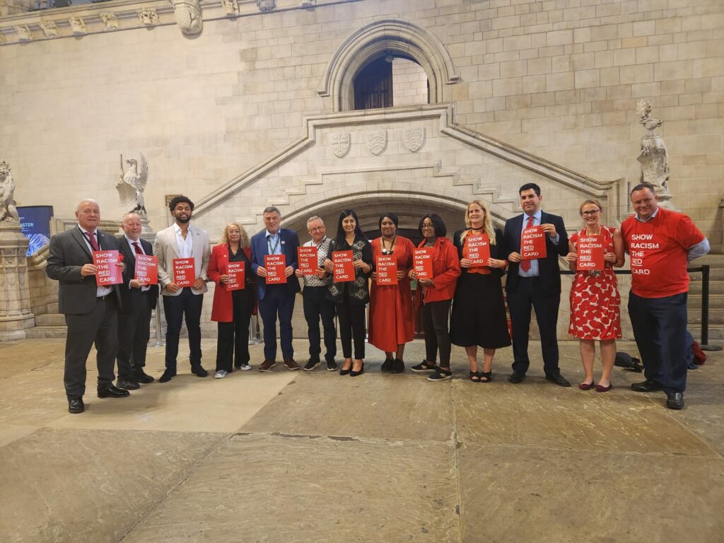 Bell standing with representatives from Show Racism the Red Card. They are holding up red signs that also display the organisation's name. 